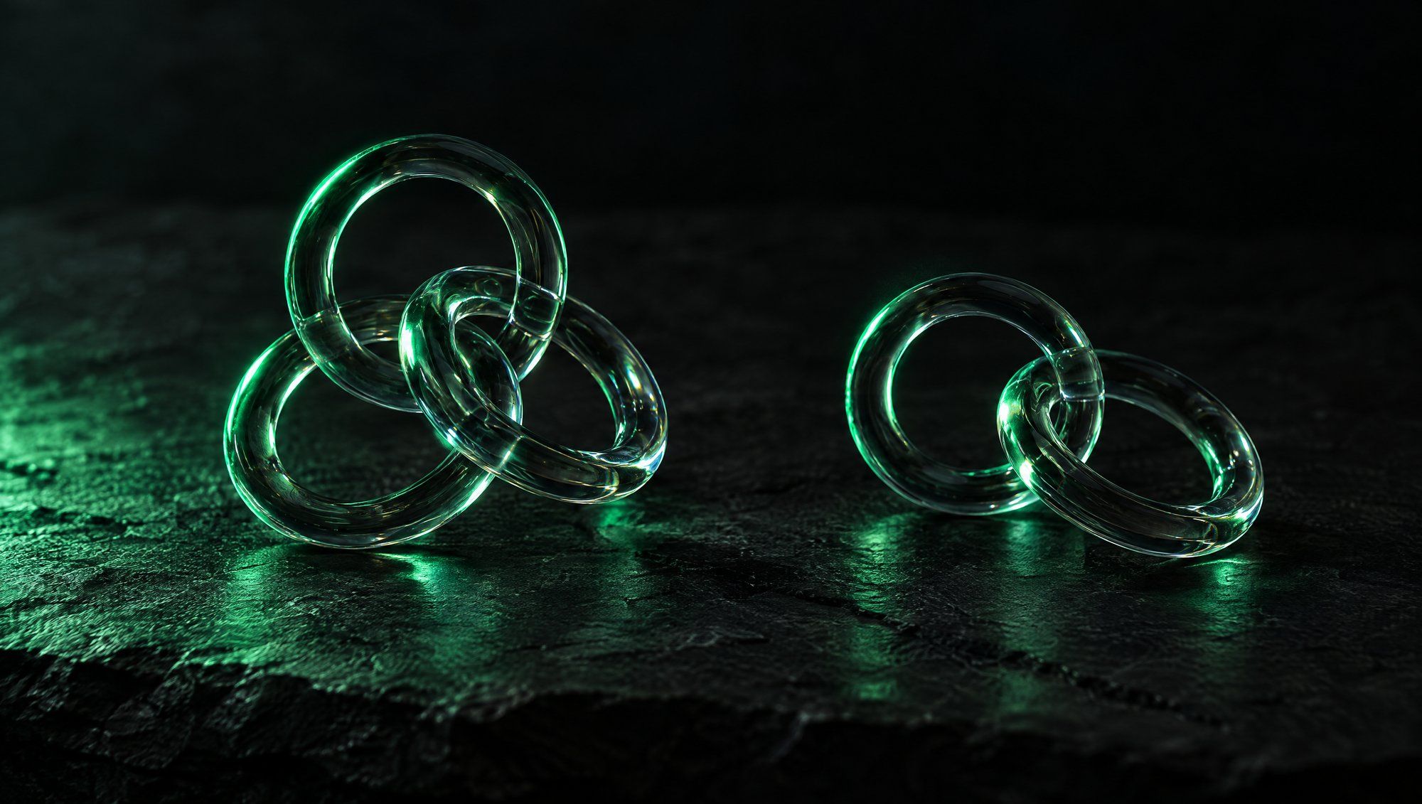 Two clear glass ring sculptures on dark slate, one made of three linked rings and one of two linked rings, both edged in green light with green reflections on the stone.