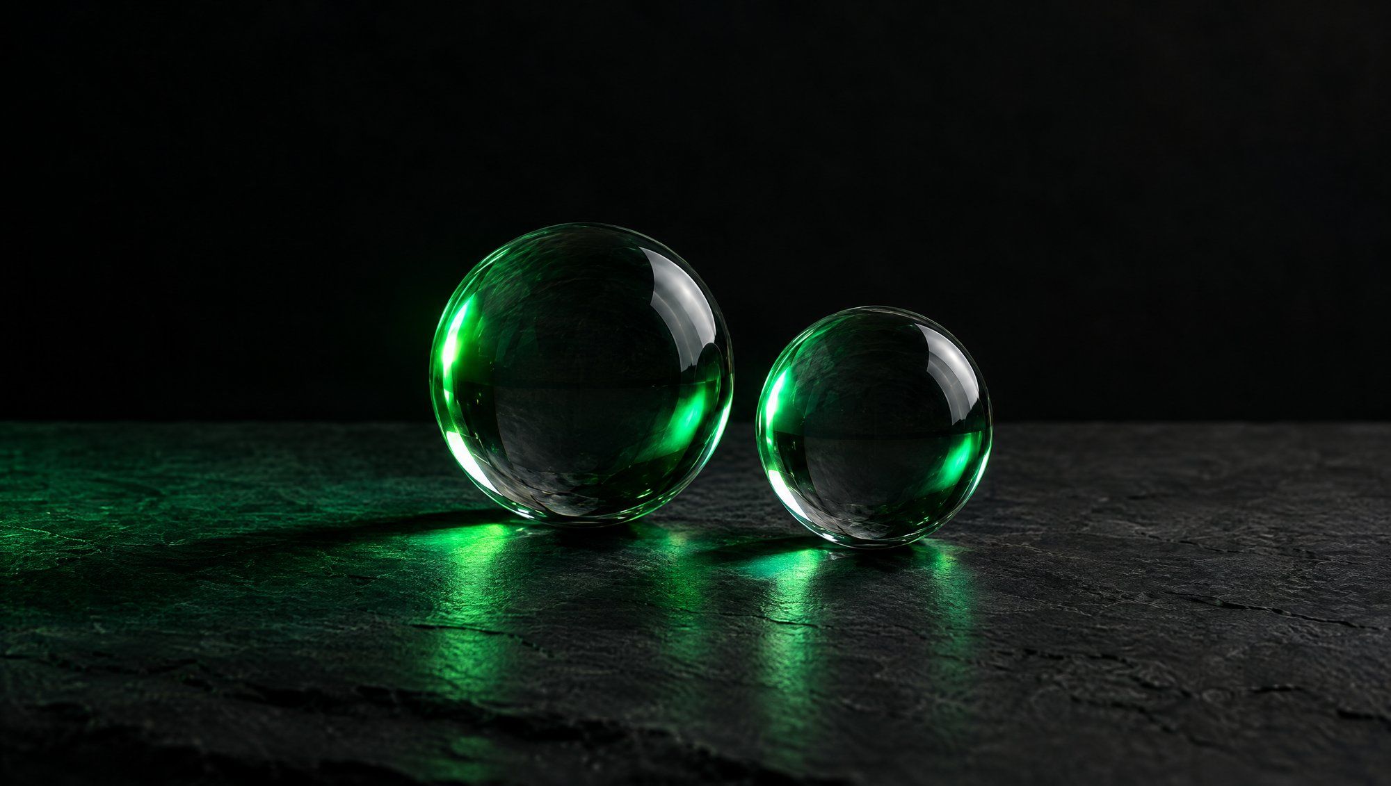 Two clear glass spheres of different sizes side by side on dark stone, lit from the side by green light.